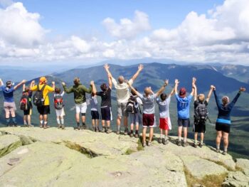 A group of people standing on a rocky ledge with their backs to the camera, arms raised towards a mountainous landscape.
