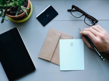 stock image of a card, a man's hand with a pencil, a notebook, business card, glasses, and a plant