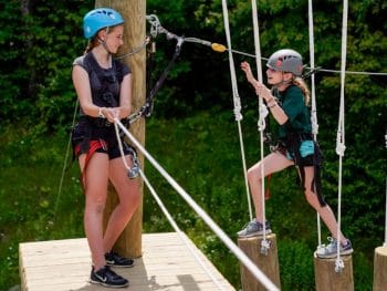 a young girl climbs on the tops of logs while another girl watches