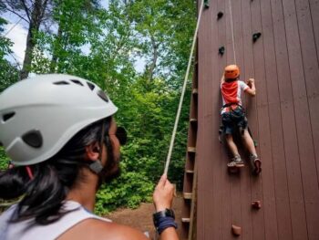 A child at summer camp wearing a helmet and harness climbs a wooden rock wall while another person, also in a helmet, stands behind them holding the rope for support.